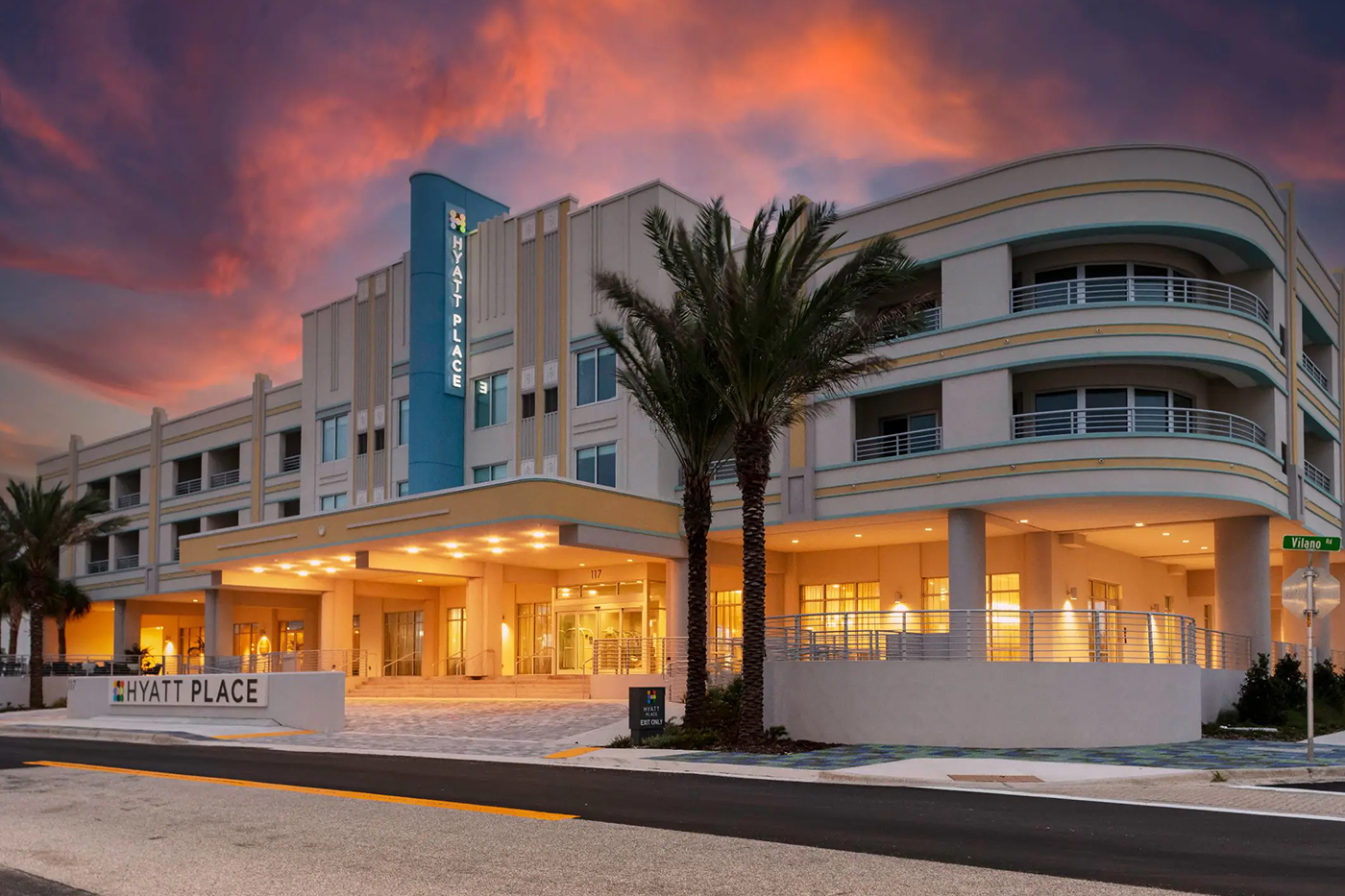 Hyatt Place St. Augustine Vilano Beach hotel exterior at sunset, Art Deco style building with palm trees and dramatic pink and orange sky, Vilano Road sign visible