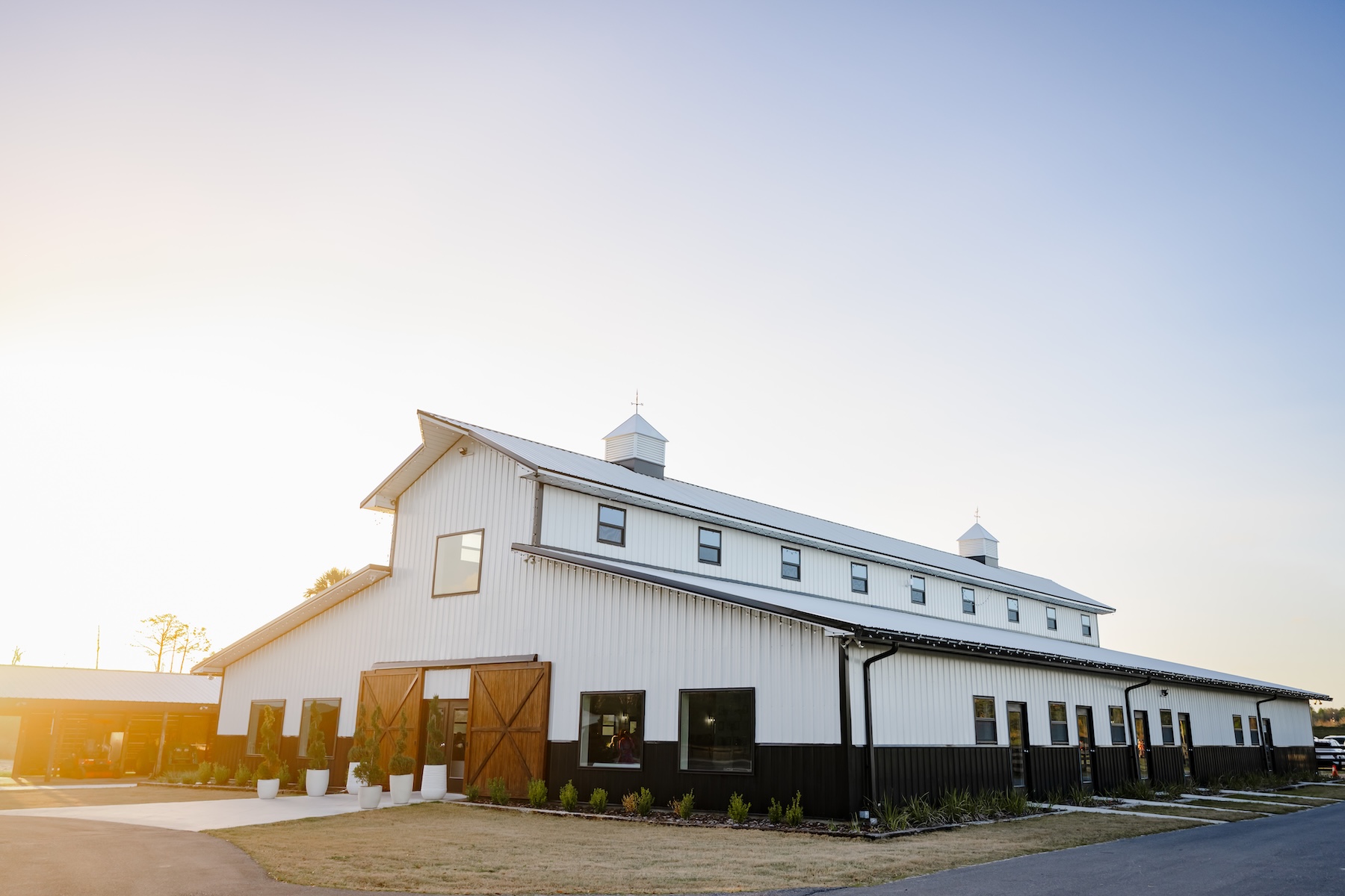 Ancient City Farmstead barn exterior at golden hour near St. Augustine FL, white vertical siding with cupolas and wooden barn doors glowing warmly in evening light