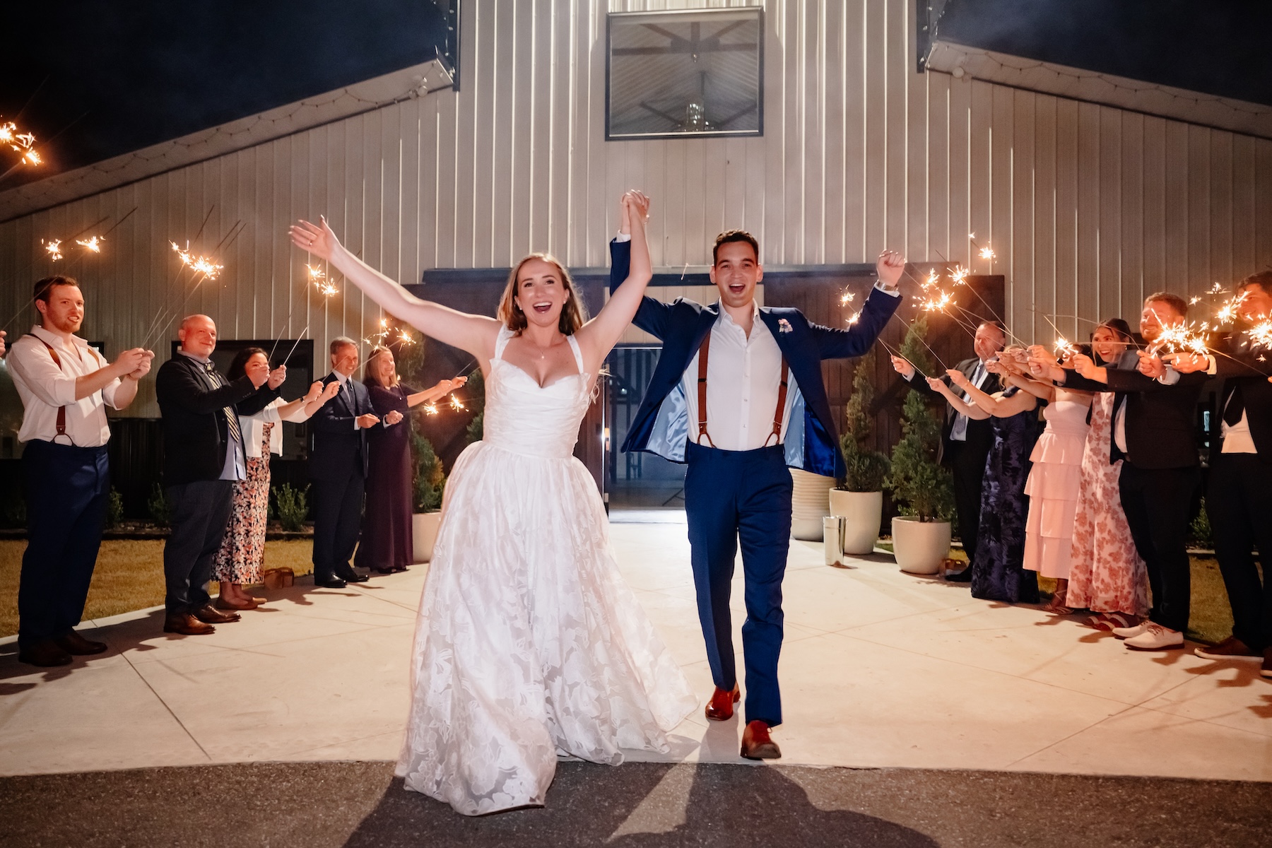 Bride and groom celebrating their sparkler exit in front of the Ancient City Farmstead barn near St. Augustine FL at night, guests lining both sides holding sparklers, barn facade lit behind them