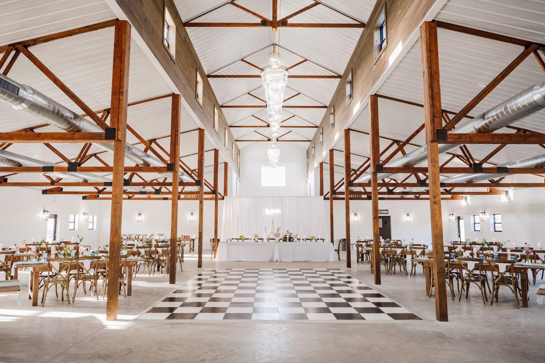 Wide interior shot of the Grand Cypress barn at Ancient City Farmstead near St. Augustine FL, vaulted ceiling with exposed wood trusses and chandelier, checkered dance floor, farm tables and cross-back chairs set for a wedding reception