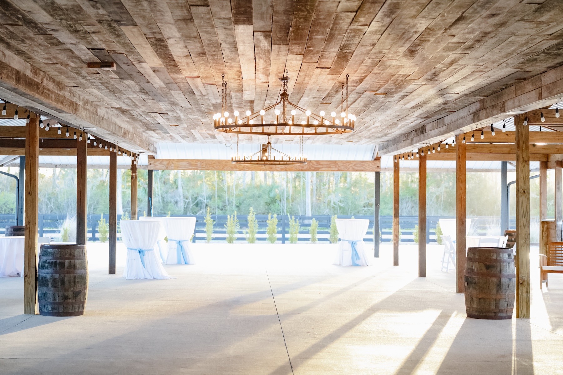 Outdoor covered pavilion at Ancient City Farmstead near St. Augustine FL, reclaimed wood plank ceiling with wagon wheel chandeliers, white cocktail tables with blue sashes, wine barrels as decor, open sides facing the treeline