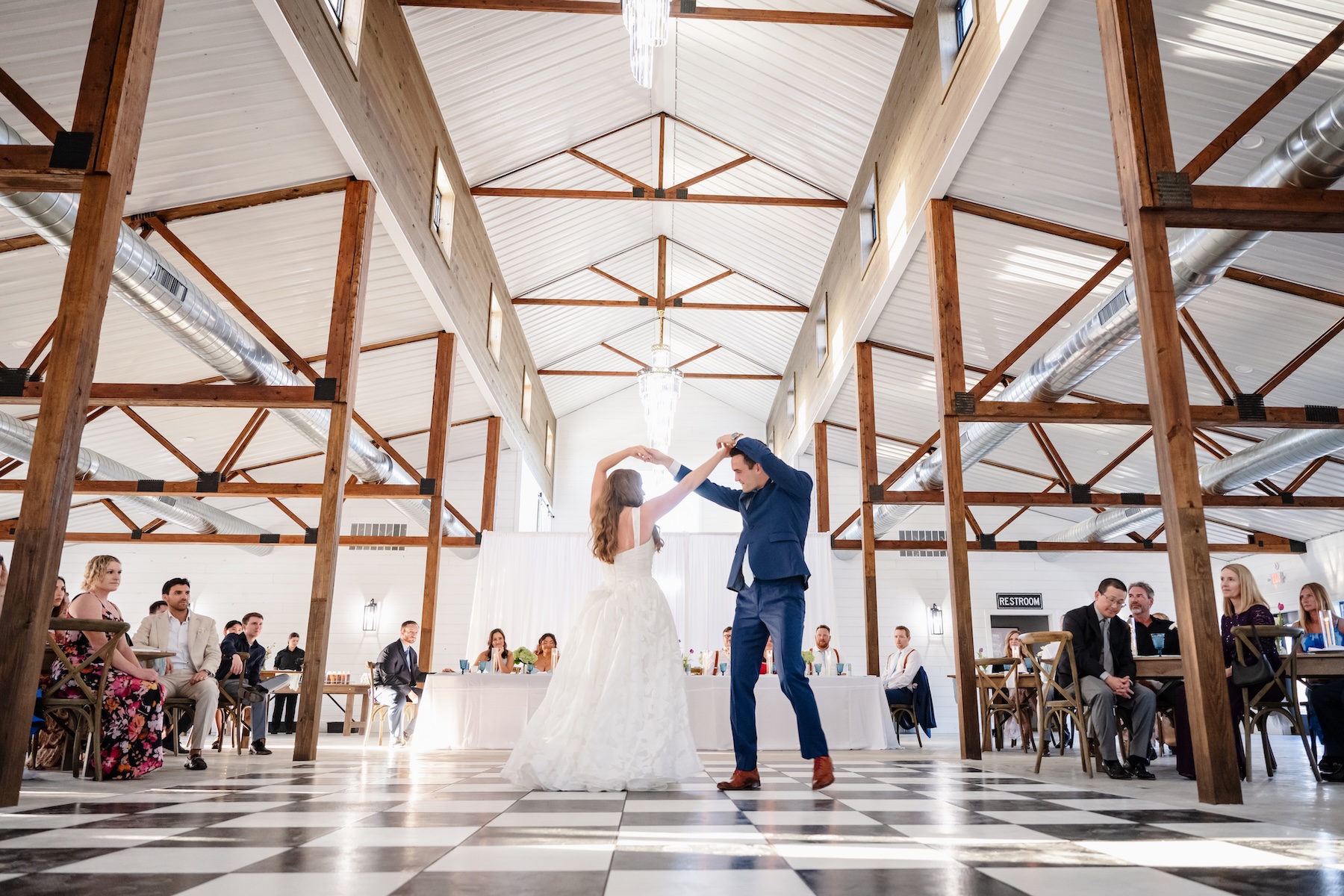 Bride and groom sharing their first dance on the checkered dance floor of the Grand Cypress barn at Ancient City Farmstead near St. Augustine FL, chandelier above, exposed wood trusses, guests watching from farm tables on both sides