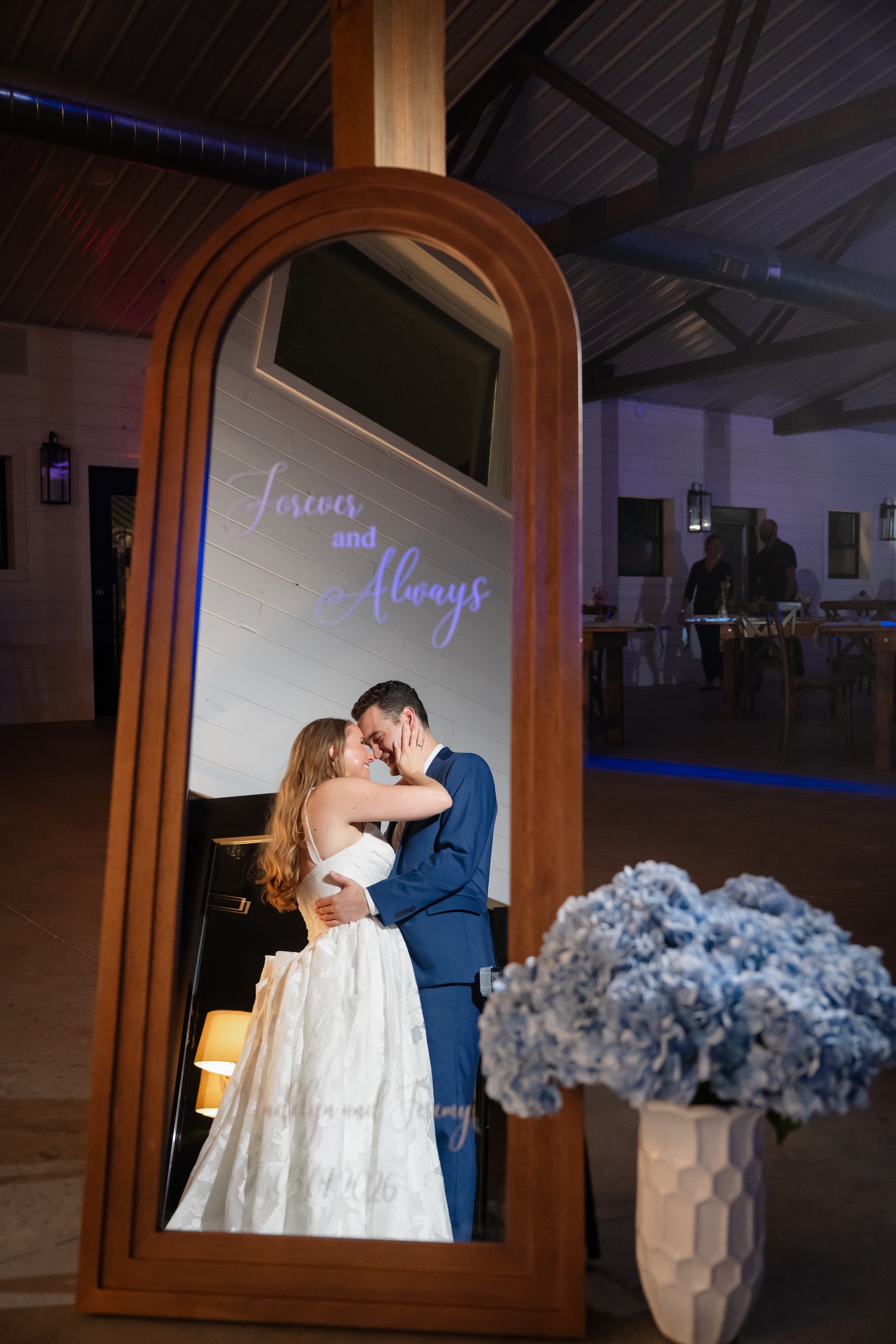 Bride and groom reflected in the Footloose Entertainment photo booth at Ancient City Farmstead near St. Augustine FL, Forever and Always displayed on screen, blue uplighting illuminating the barn interior