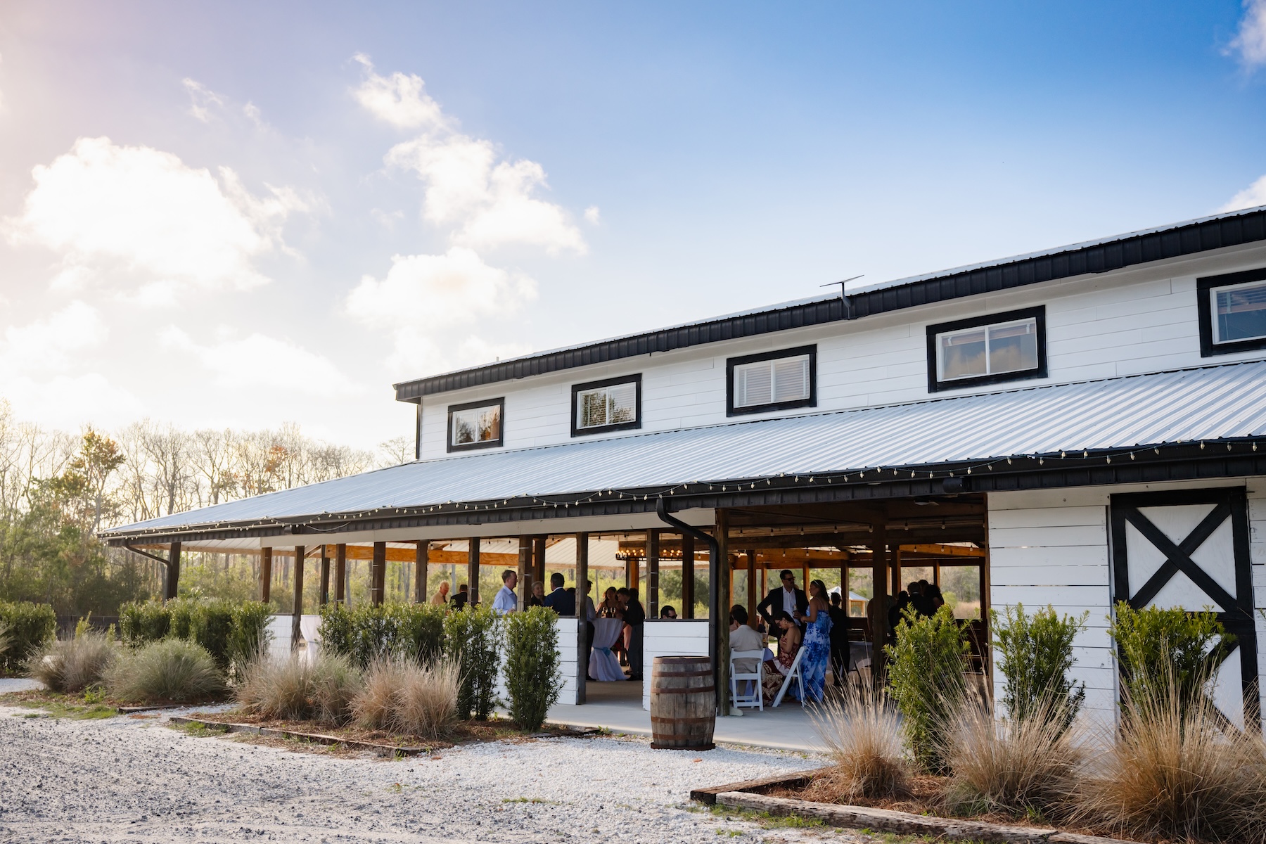 Ancient City Farmstead barn exterior near St. Augustine FL with wedding cocktail hour guests mingling under the covered porch, wine barrel planter and lush landscaping, blue sky and clouds behind