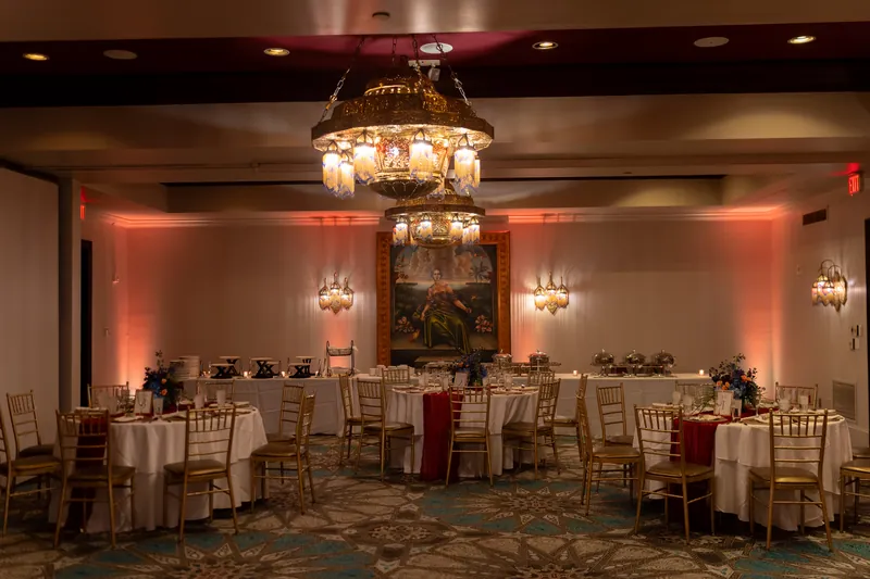 Flagler Ballroom at Casa Monica Hotel in St. Augustine FL set for a wedding reception, ornate chandelier, pink uplighting, gold chiavari chairs, round tables with white linens and red napkins, historic painting on the wall