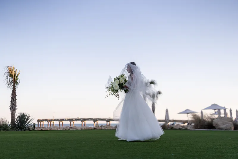 Bride in white gown standing on the waterfront lawn at Embassy Suites St. Augustine FL at dusk, pier and Intracoastal water in the background, palm trees and resort grounds
