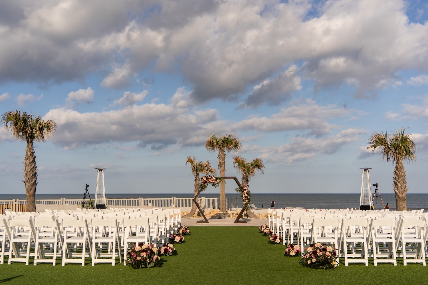 Wide view of outdoor ceremony setup at Embassy Suites St. Augustine, white chairs in rows, floral arch, palm trees and ocean view behind the altar