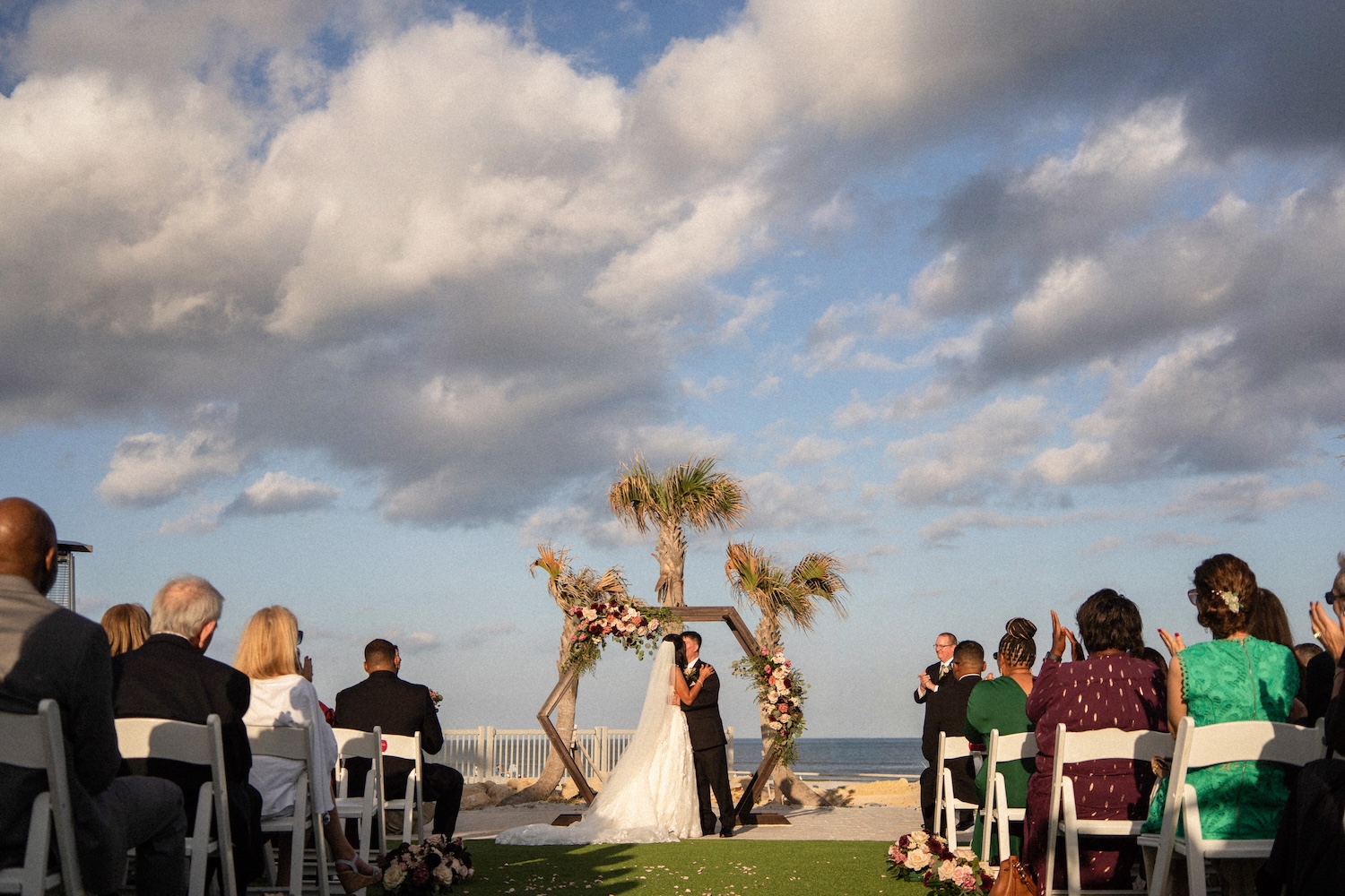 Wedding ceremony at Embassy Suites St. Augustine from guest perspective, couple at altar with floral arch, ocean view, guests seated on white chairs