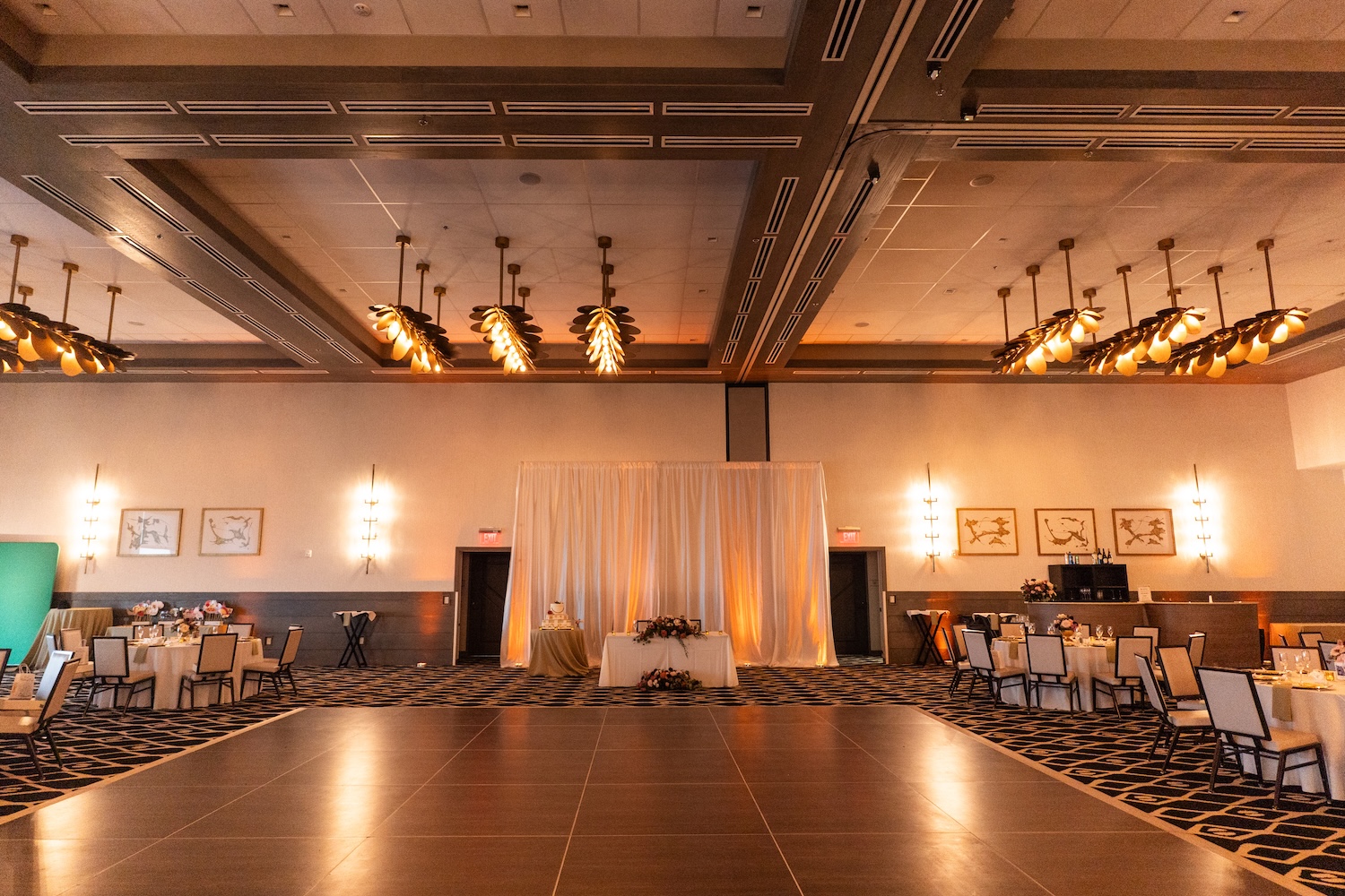 Embassy Suites St. Augustine ballroom reception setup, centered dance floor with draped backdrop, warm amber uplighting, tables arranged around the room