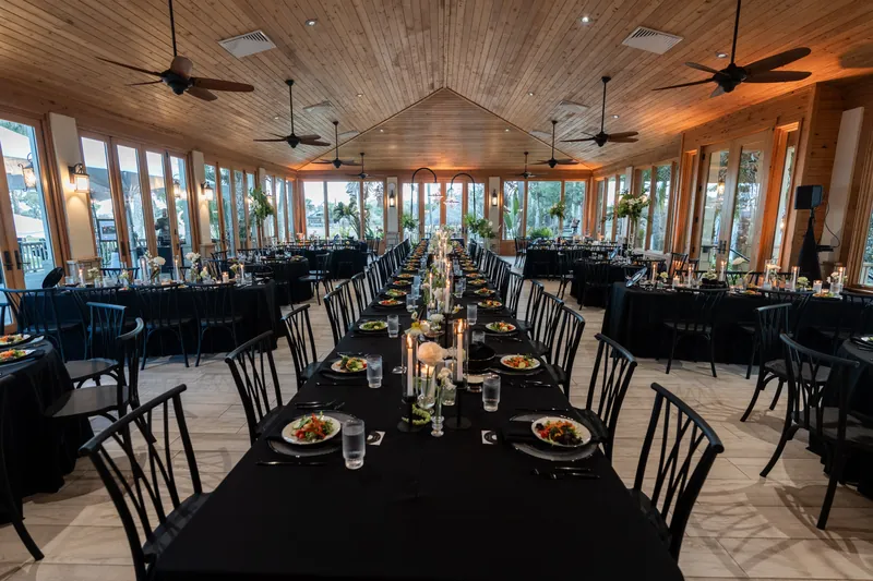Elegant wedding reception inside the pavilion at Fountain of Youth Archaeological Park in St. Augustine FL, long farm tables with black linens, tall white floral and candle centerpieces, wood ceiling with ceiling fans and floor-to-ceiling windows overlooking the grounds