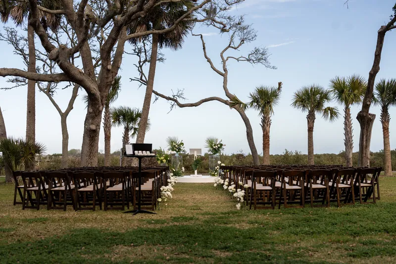 Outdoor wedding ceremony setup at the Fountain of Youth Archaeological Park in St. Augustine FL, rows of brown folding chairs on the lawn under ancient live oaks and palm trees, white floral aisle, arbor visible in the distance