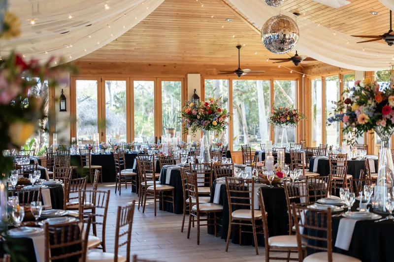 Festive wedding reception inside the Fountain of Youth Archaeological Park pavilion in St. Augustine FL, round tables with chiavari chairs and colorful floral centerpieces, disco ball and white ceiling draping, floor-to-ceiling windows with garden views