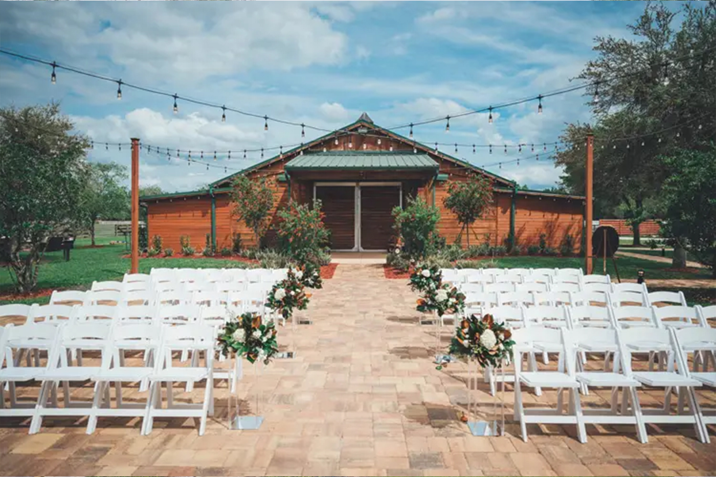 Kelly Farm Events outdoor wedding ceremony setup near St. Augustine FL, white chairs on brick patio with floral arrangements lining the aisle, wood barn and string lights in the background