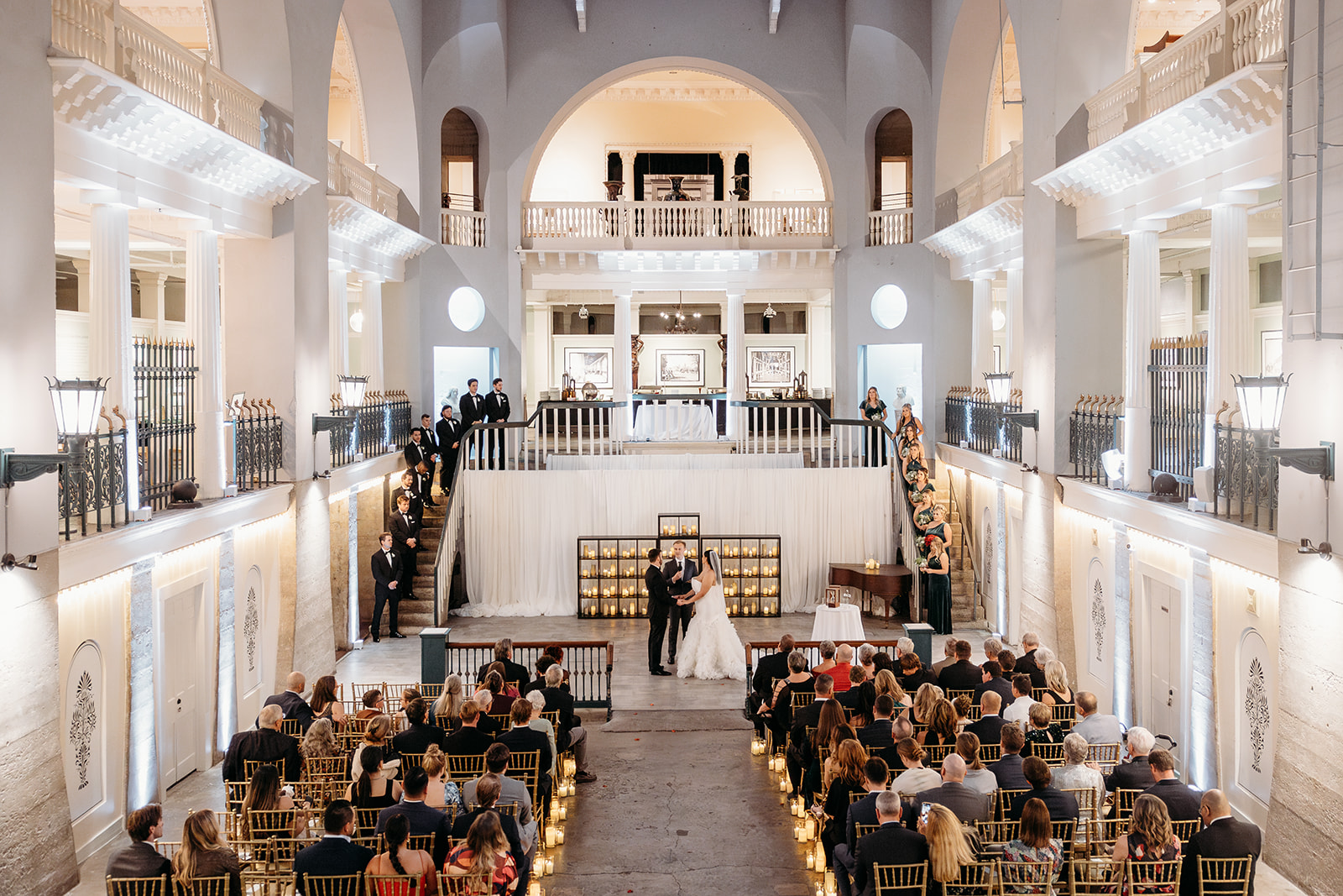 Wedding ceremony in the Alcazar Court at the Lightner Museum St. Augustine FL — guests seated on the main floor with couple at the altar, dual staircases and balconies above — Footloose Entertainment wedding DJ