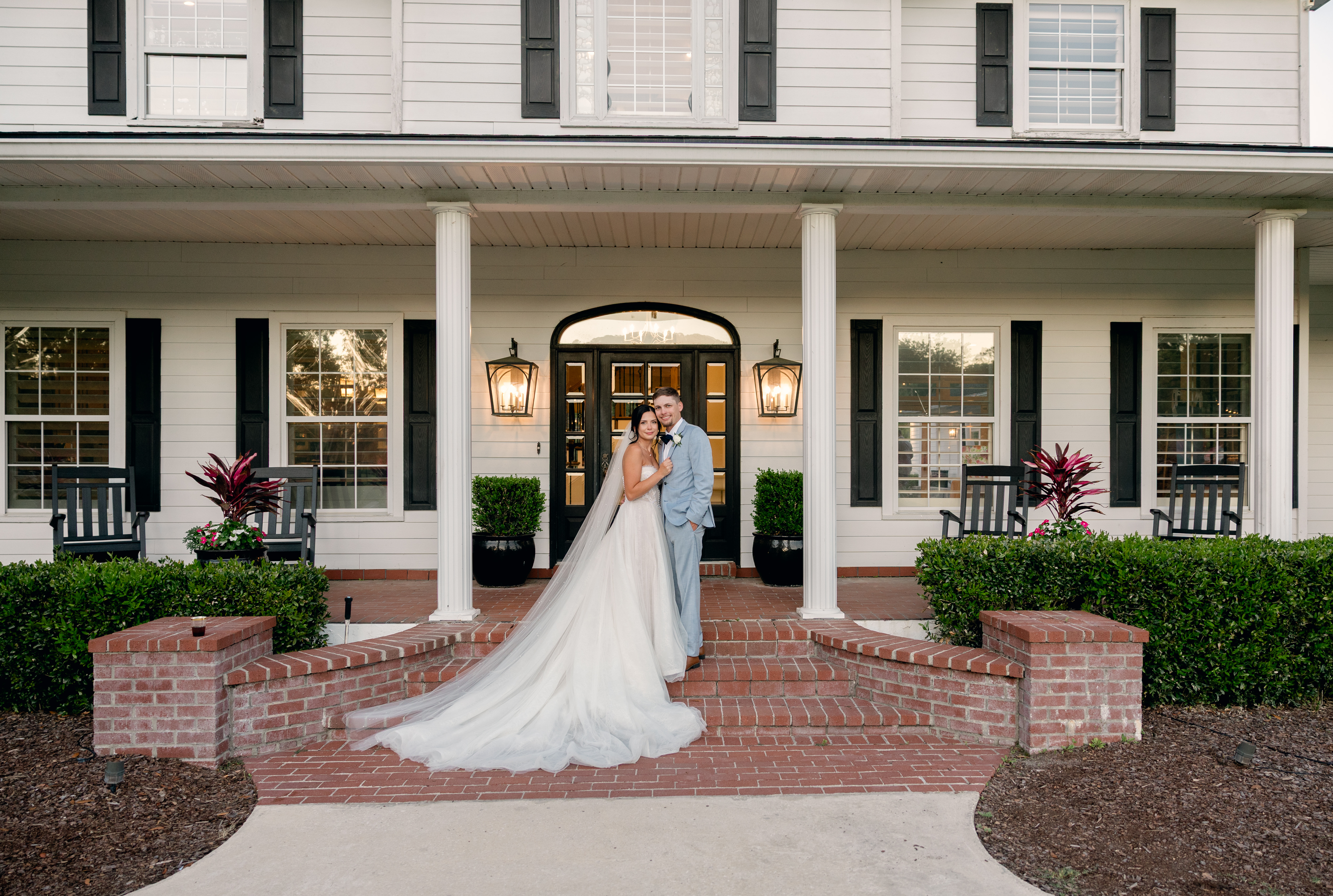 Bride and groom posing on the front porch brick steps of The Manor at 12 Oaks in Green Cove Springs FL, white columns, black arched door and lantern sconces behind them