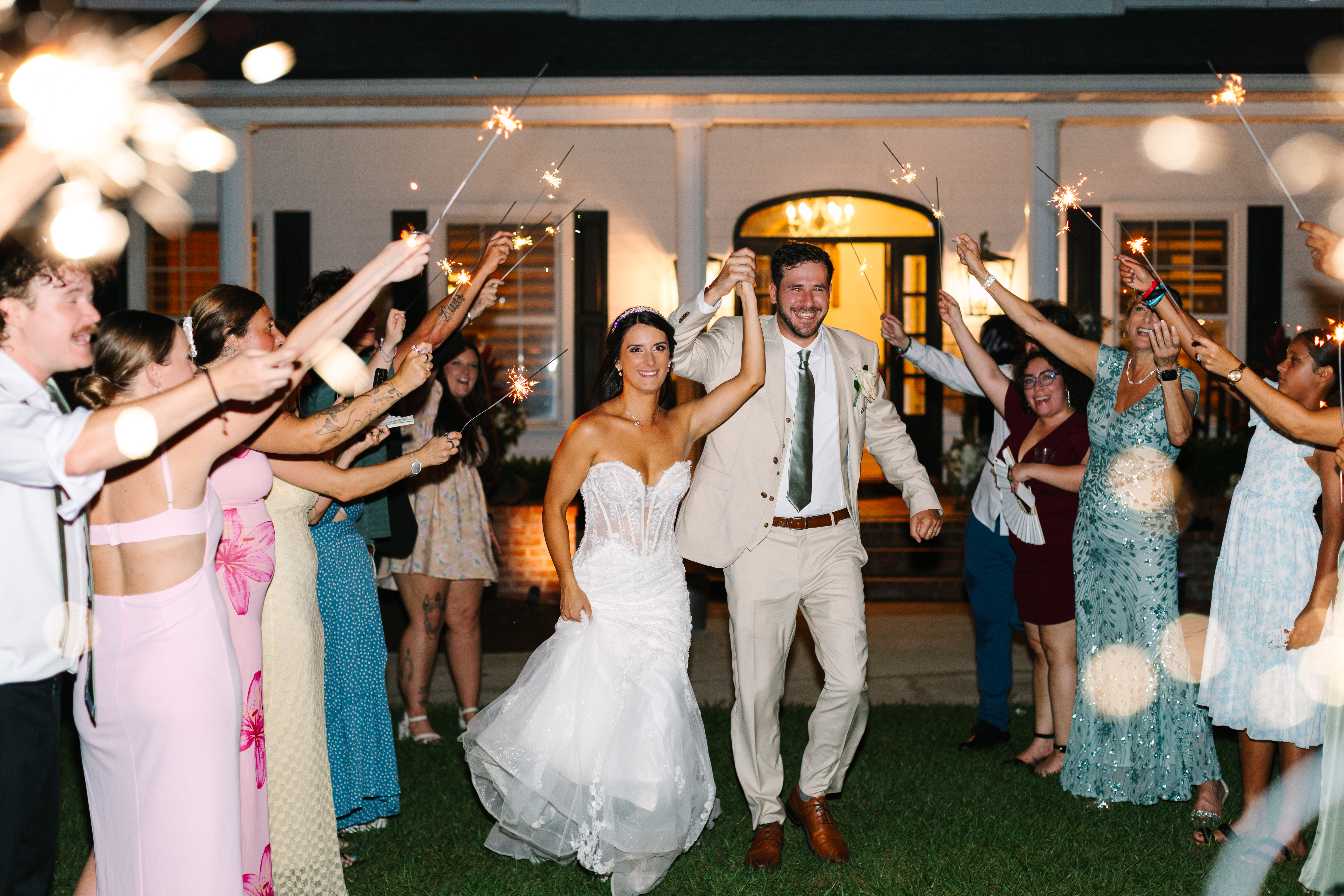Bride and groom running through a sparkler exit tunnel at The Manor at 12 Oaks at night, the Manor house with white columns and arched door glowing warmly behind them