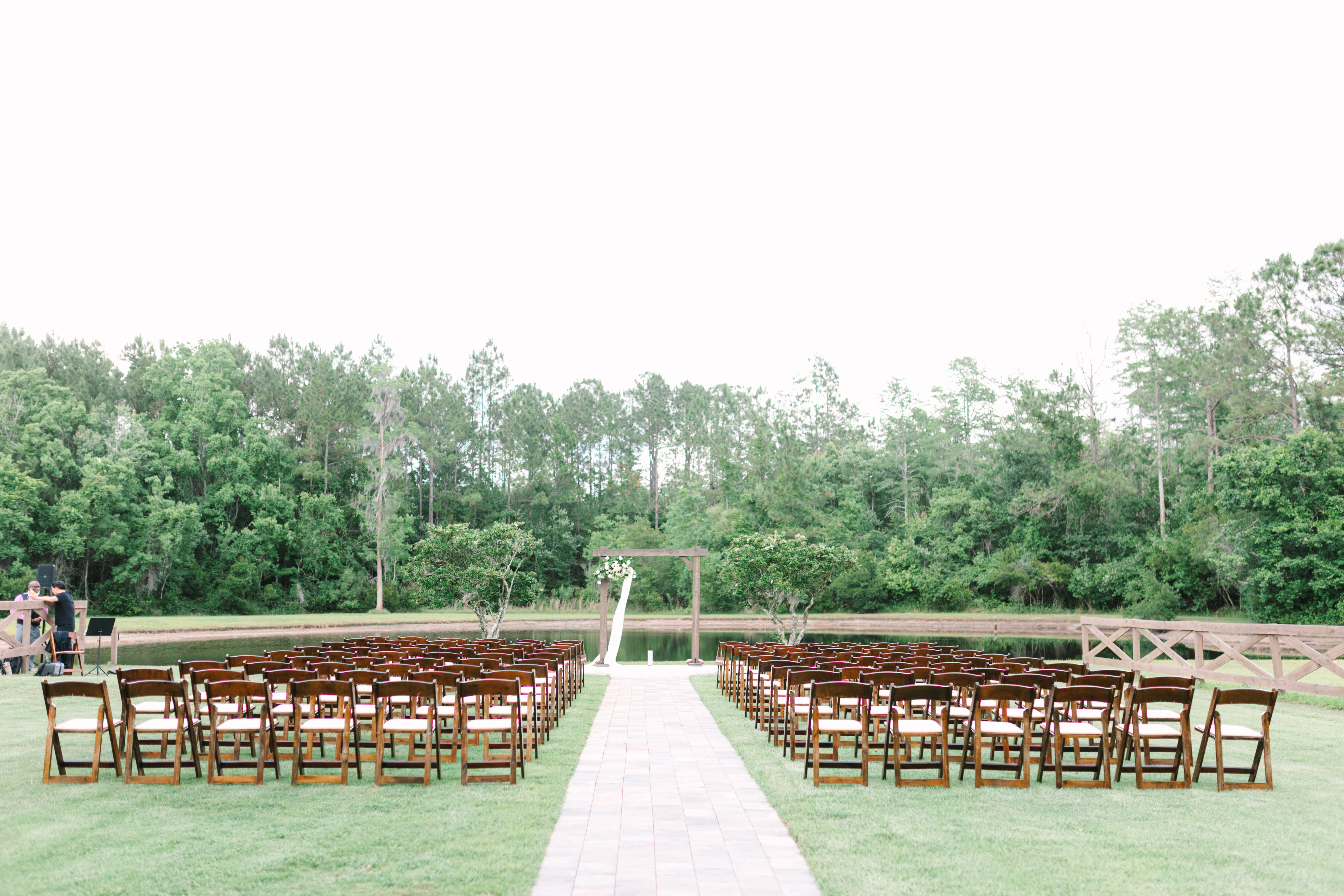 Wide view of outdoor wedding ceremony setup at The Manor at 12 Oaks Green Cove Springs FL, rows of wooden folding chairs on green lawn, wooden arbor with white florals at the end, pond and treeline behind