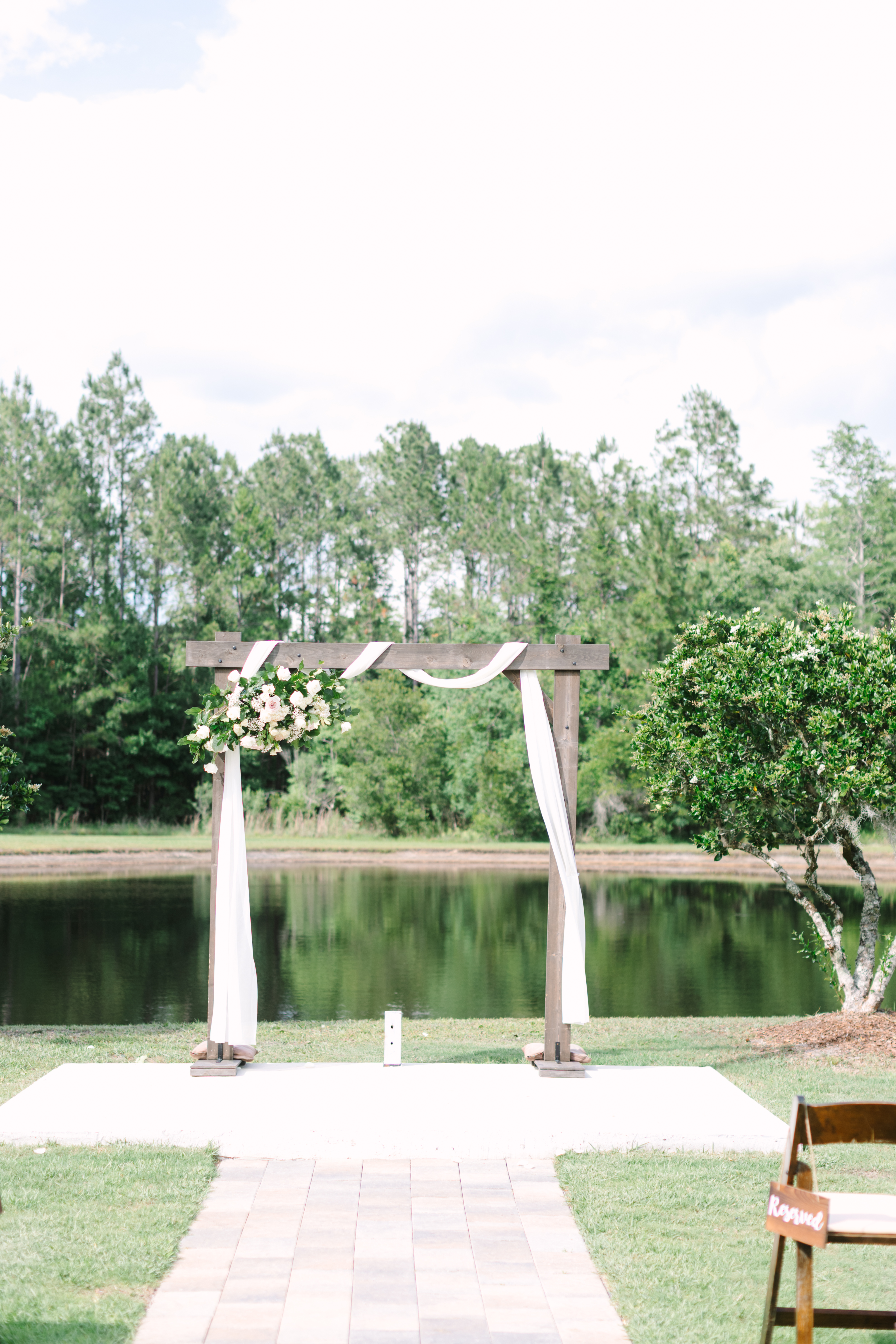 Wooden ceremony arch draped with white fabric and white florals at The Manor at 12 Oaks with the pond and lush treeline perfectly reflected in the water behind it