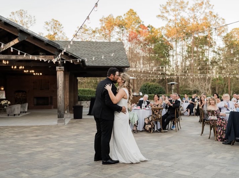 Wide shot of outdoor wedding reception on the stone patio at The Manor at 12 Oaks Green Cove Springs FL, guests seated at round tables, string lights, barn pavilion structure and fall foliage in background