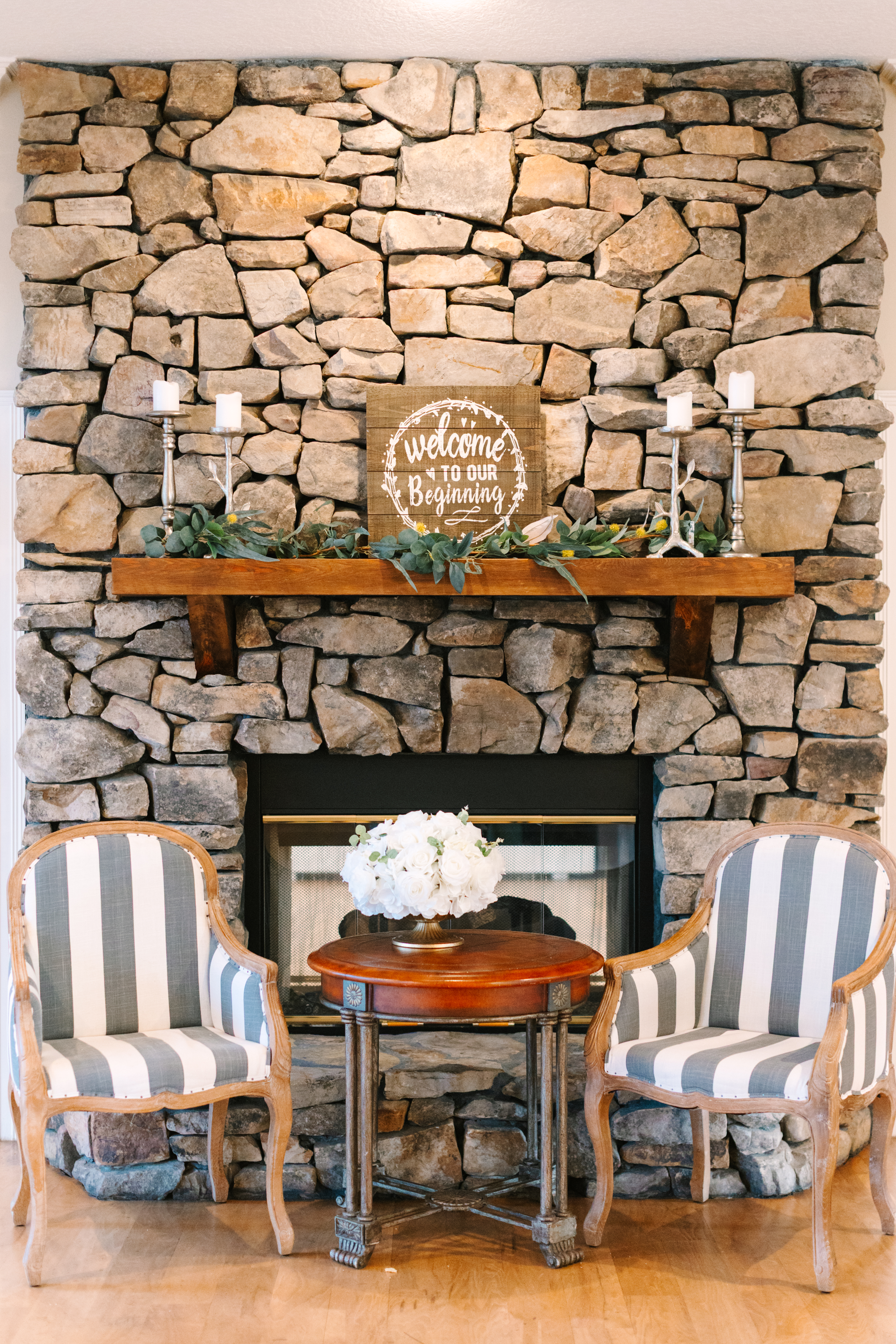 Large stone fireplace lounge inside The Manor at 12 Oaks Green Cove Springs FL, two striped arm chairs flanking a round wooden table with white hydrangea arrangement