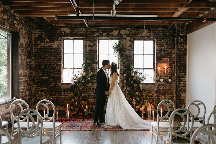Wedding ceremony inside The Lark event venue in downtown Jacksonville FL, bride and groom at a floral arch altar in an exposed brick and wood-beam interior, candles and vintage rugs