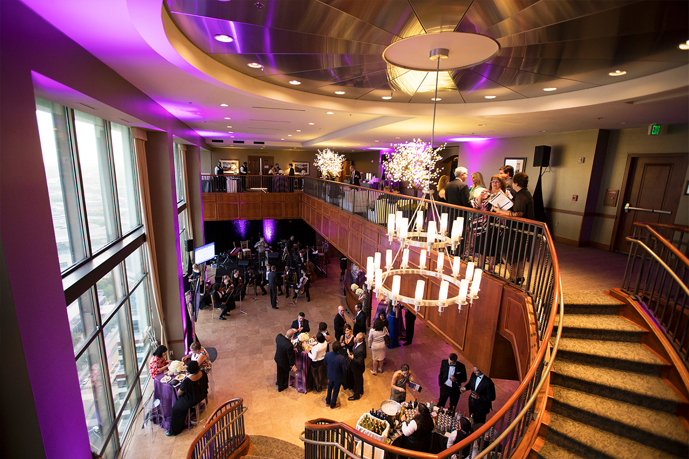 The River Club event space interior in downtown Jacksonville FL, two-level reception with curved staircase and chandelier, purple uplighting, guests socializing on 34th floor with city views