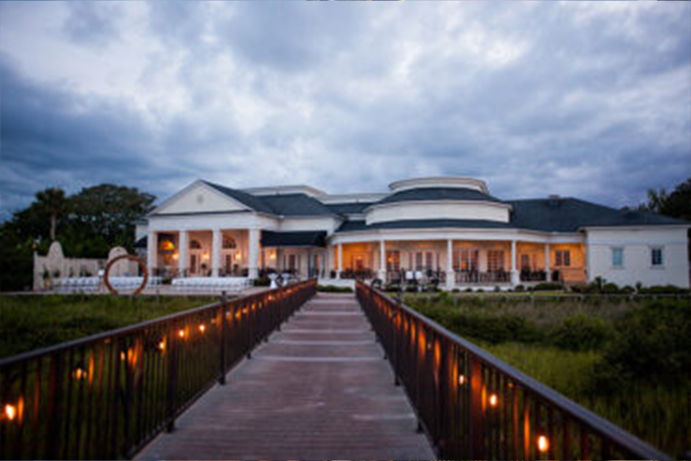 The River House waterfront wedding venue in downtown St. Augustine FL, illuminated white mansion viewed from a lit footbridge over the marsh at dusk