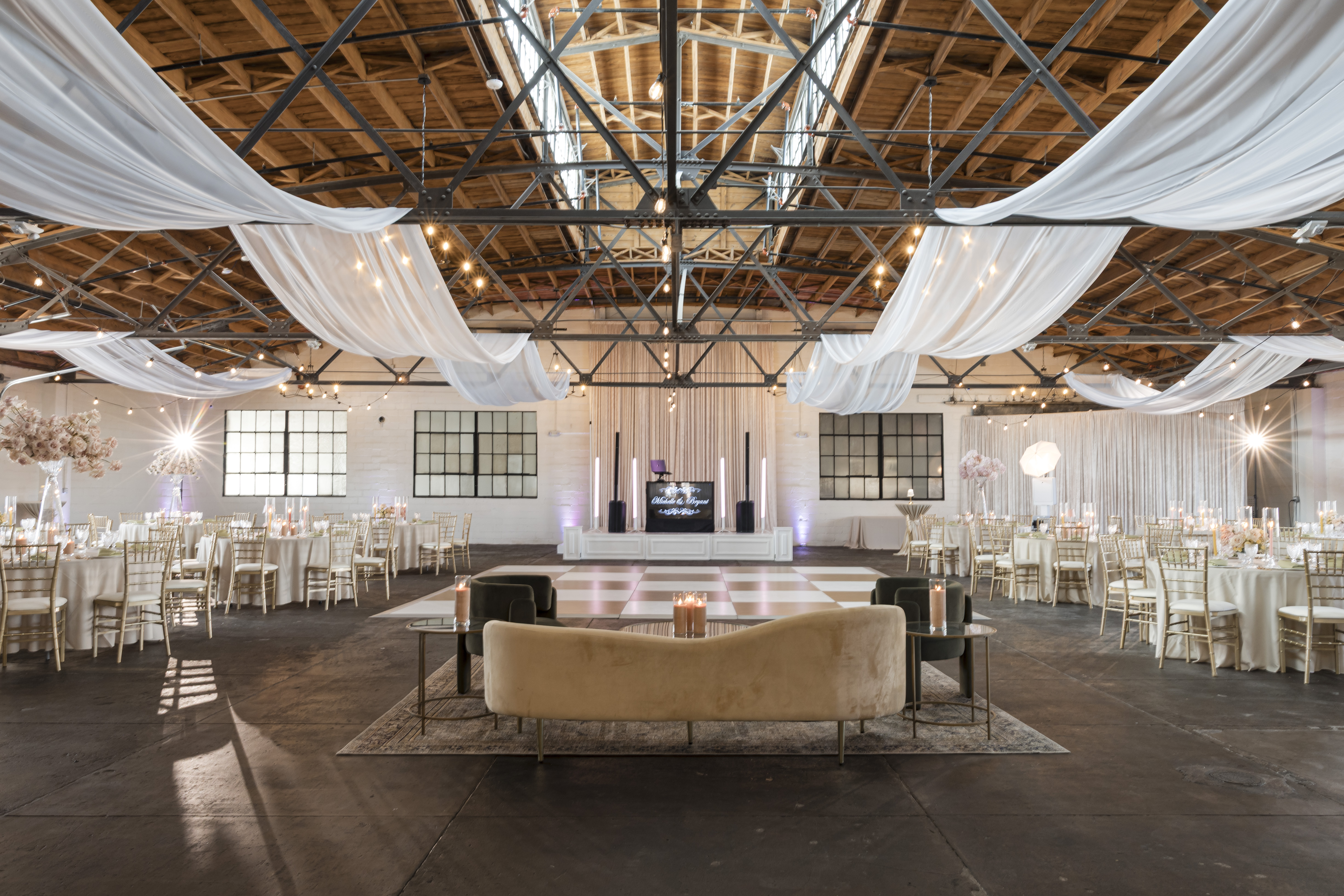 Full ballroom at The Venue Hastings showing checkered dance floor, DJ booth with monogram display, lounge seating, and industrial truss ceiling with white draping