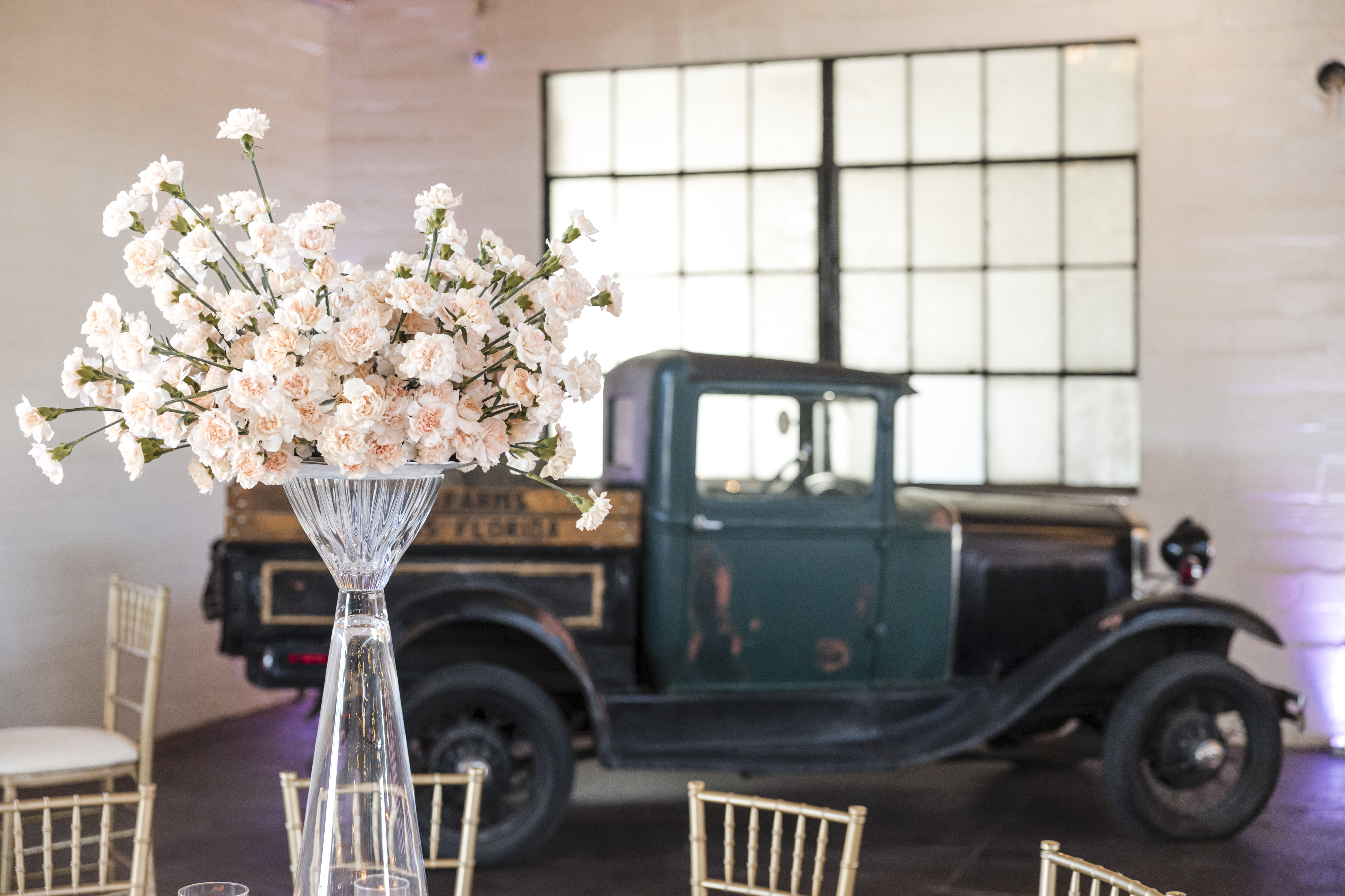 Vintage antique truck parked inside The Venue Hastings Great Room with tall blush floral arrangement in crystal vase and gold chiavari chairs