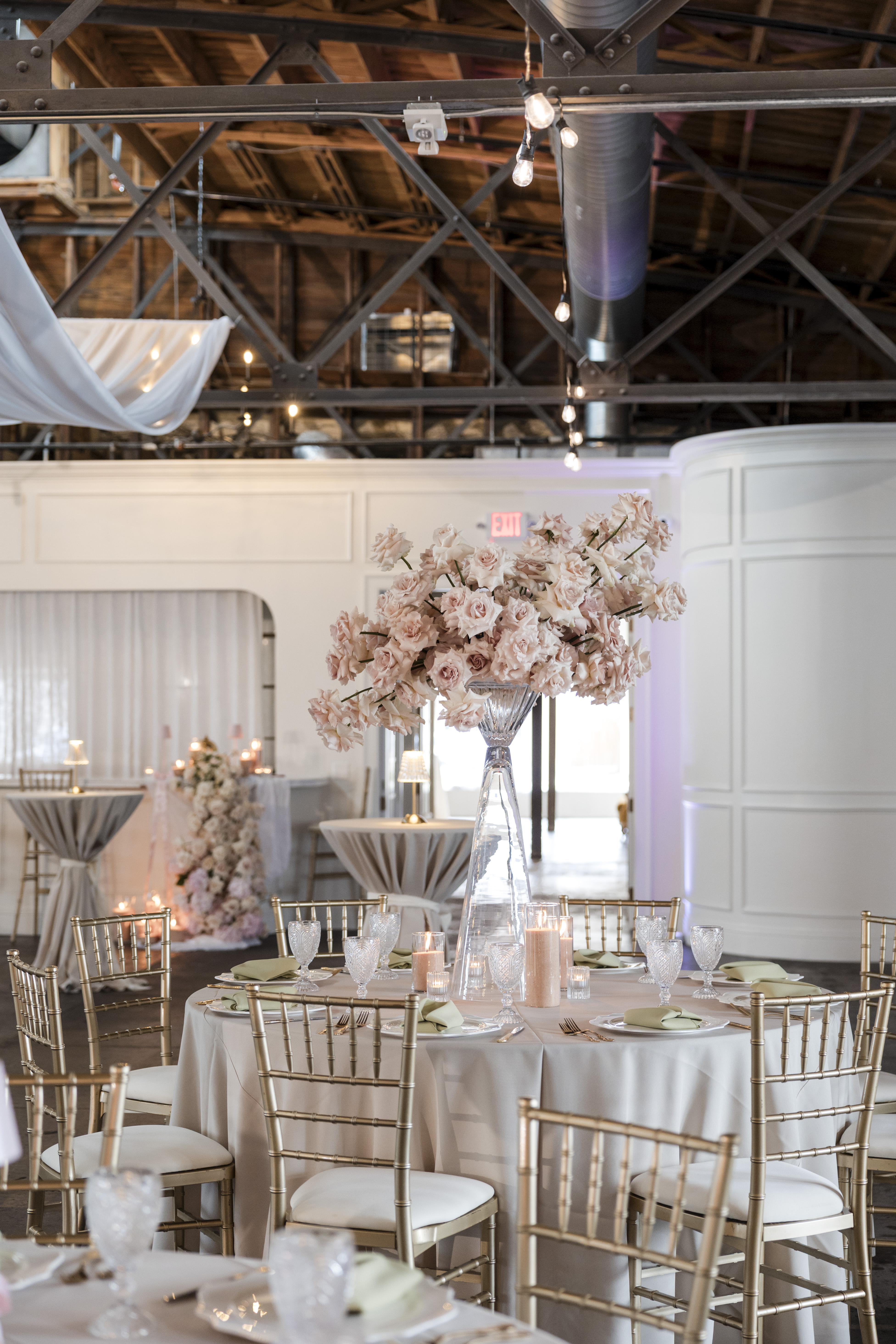 Reception table setup at The Venue Hastings with tall blush rose centerpiece in crystal vase, gold chiavari chairs, candles, and industrial truss ceiling with string lights