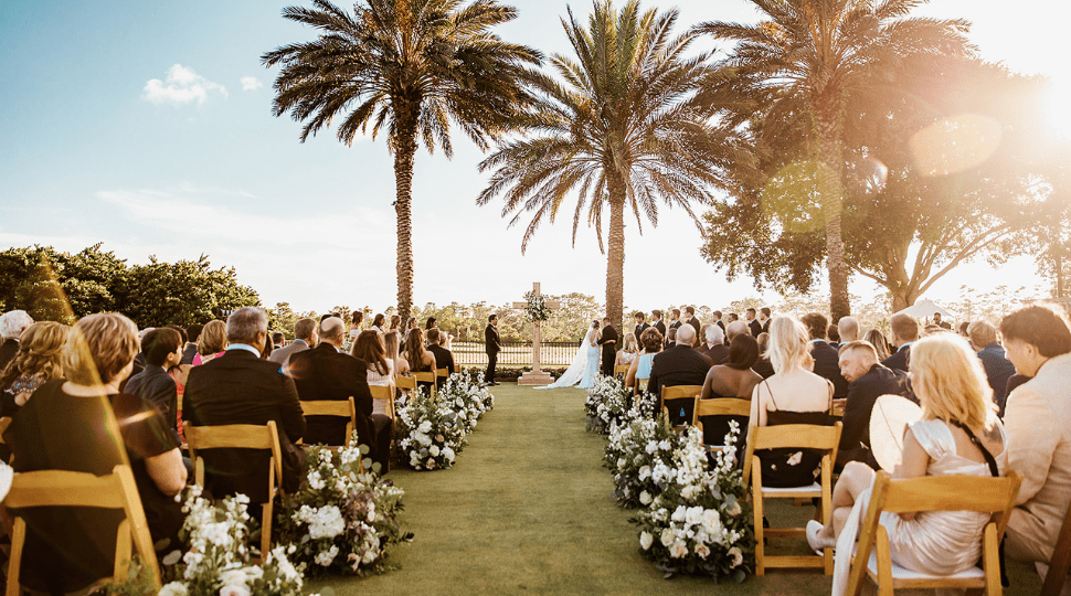 Outdoor wedding ceremony on the event lawn at TPC Sawgrass in Ponte Vedra Beach FL, guests seated in wooden chairs lining a white floral aisle, tall palm trees and golden afternoon sun
