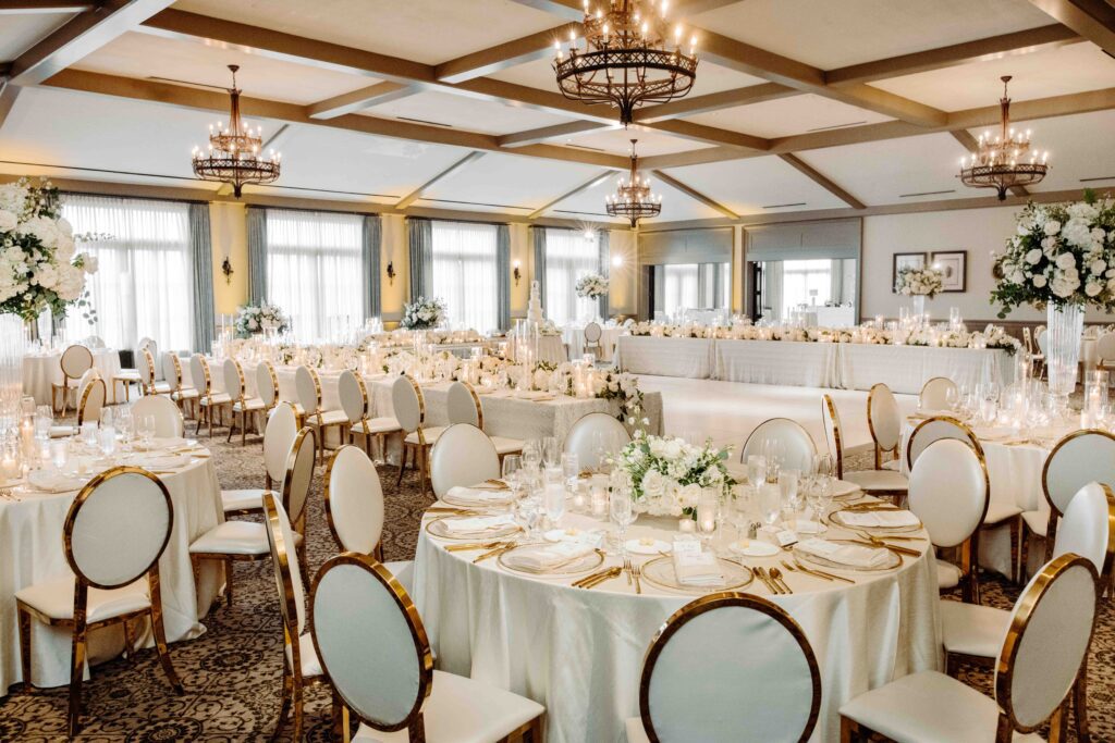 Elegant wedding reception in the TPC Sawgrass clubhouse banquet room in Ponte Vedra Beach FL, white linens, gold oval-back chairs, iron chandeliers, white floral centerpieces, coffered ceiling