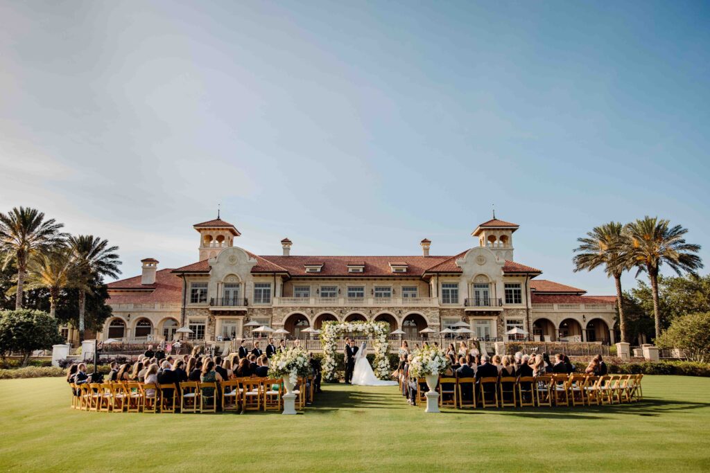 Wedding ceremony on the event lawn at TPC Sawgrass in Ponte Vedra Beach FL, bride and groom at a floral arch altar with the iconic Mediterranean Revival clubhouse behind them, guests seated on the manicured green